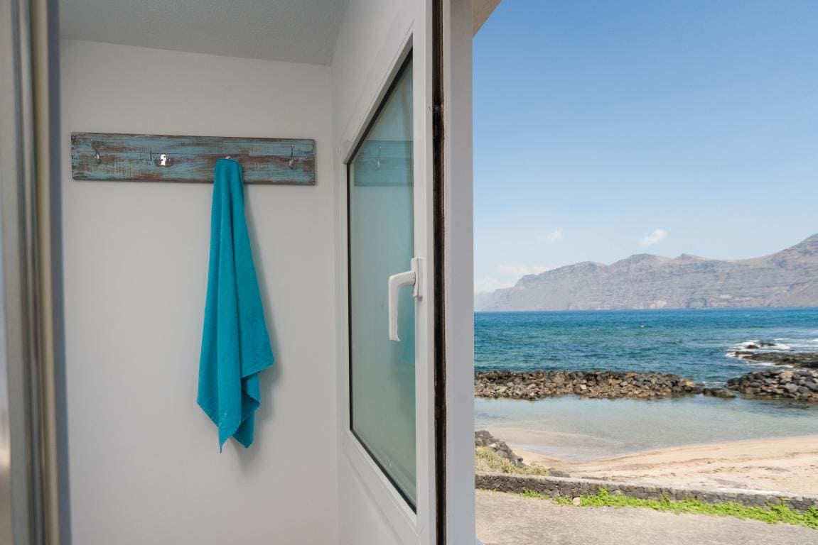 A bright, minimalistic area features a turquoise towel hanging on a rustic wooden hook. A large window provides a view of the ocean, showcasing the sandy beach and rocky coastline with distant mountains on the horizon.