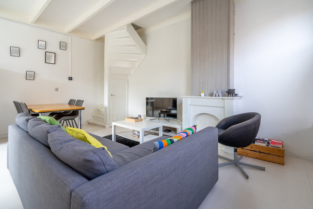 A modern living space features a comfortable gray sofa with a colorful throw, a sleek armchair, and a minimalist dining table. A television rests on a coffee table against a light wall, with a spiral staircase visible in the background leading to the upper level.