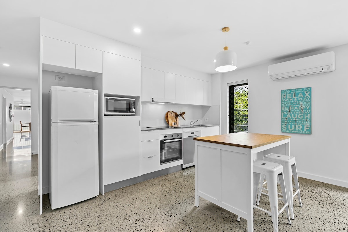 The modern kitchen is designed with sleek cabinetry and a spacious island featuring a wooden countertop. Stainless steel appliances, including a refrigerator and oven, are integrated, while natural light enters through a window. A framed wall art piece adds a touch of color to the neutral palette.
