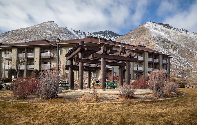 A charming outdoor area features a wooden pergola surrounded by lush landscaping. The expansive building sits in the background, flanked by rolling hills. The scene is complemented by soft blue skies and scattered clouds.