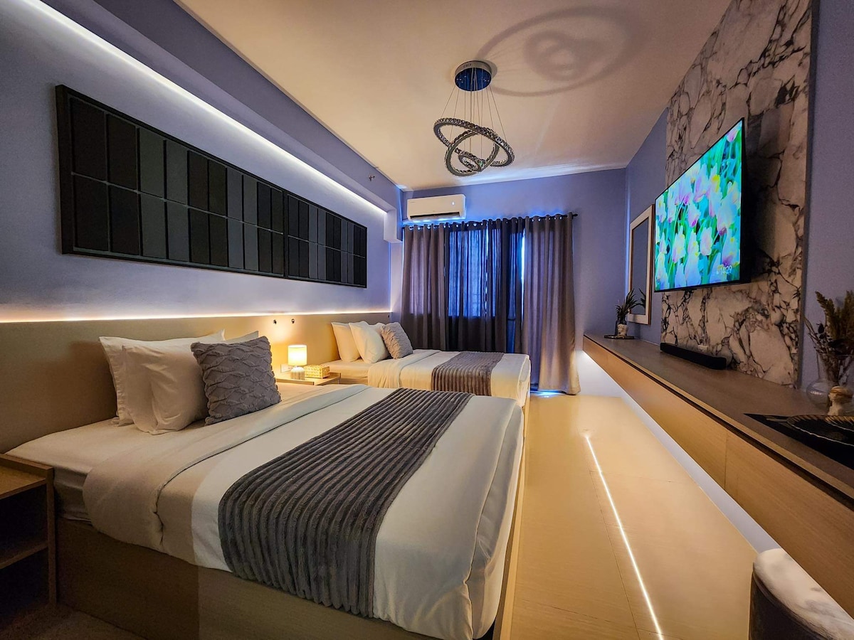 A well-lit sleeping area features a combination of two spacious beds adorned with neutral bedding. Modern lighting accents the room, complemented by a sleek wall-mounted television. Window coverings soften natural light, while stylish decor adds a contemporary touch to the space.