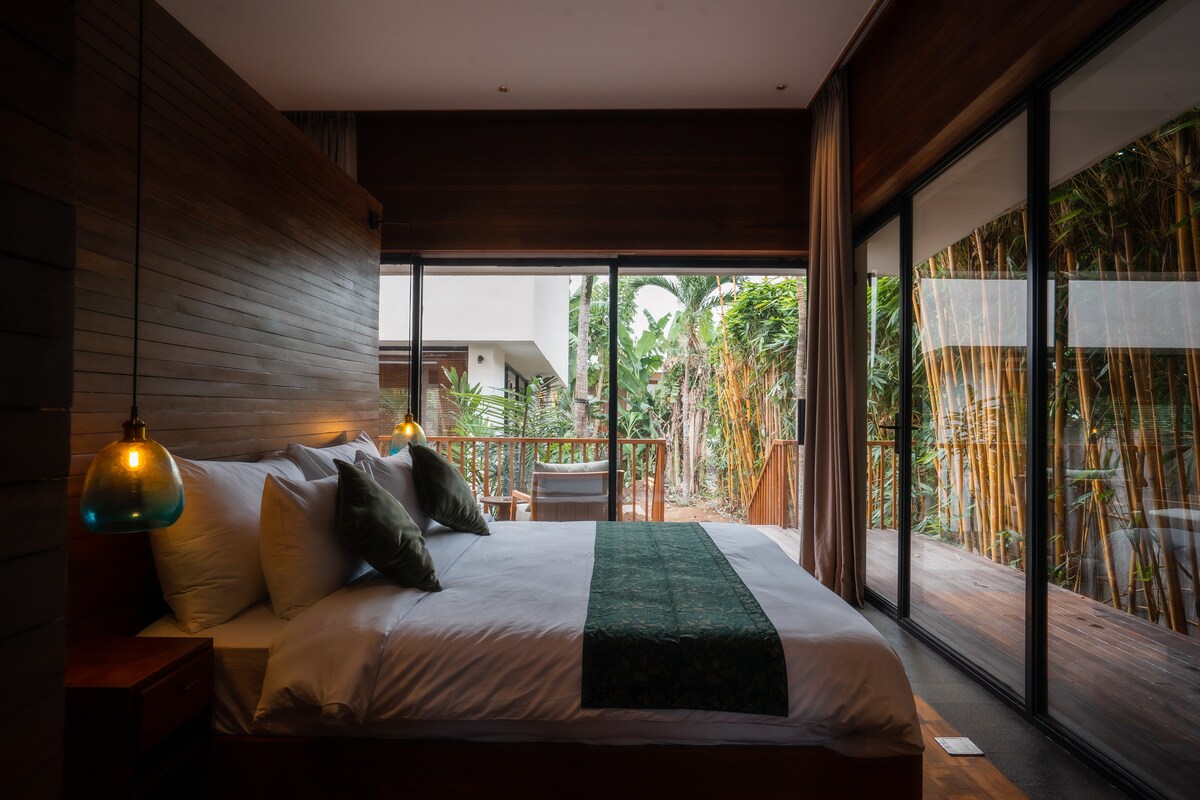 A spacious bedroom is bathed in natural light, featuring a large bed with decorative pillows and a green accent throw. Floor-to-ceiling glass doors open to a lush outdoor area, enhancing the sense of tranquility. Soft lighting is provided by stylish pendant lamps.