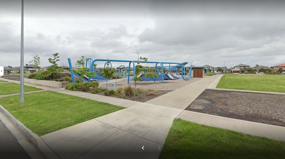A playground is visible, featuring blue play structures set against a backdrop of greenery. Paved pathways and grassy areas surround the site, providing accessible routes. Cloudy skies frame the scene, indicating a calm day in the neighborhood.