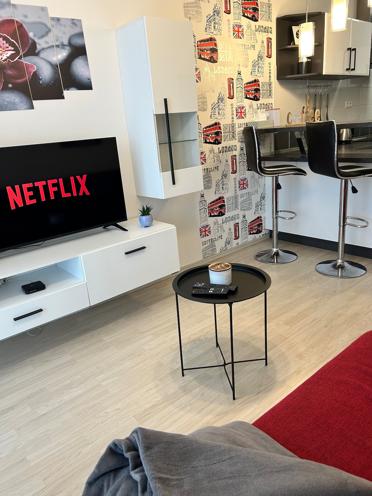 A modern living space features a sleek television displaying the Netflix logo. A small black table holds a remote control. High-backed bar stools align with a kitchen counter against a wall adorned with London-themed wallpaper.