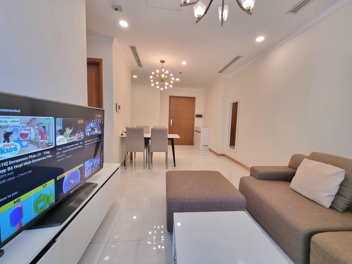 A contemporary living area features a light-colored sofa and a white entertainment unit with a large screen displaying vibrant content. A dining table for four is positioned nearby, while soft lighting from a chandelier enhances the spaciousness. The ceramic tiled floor reflects natural light.