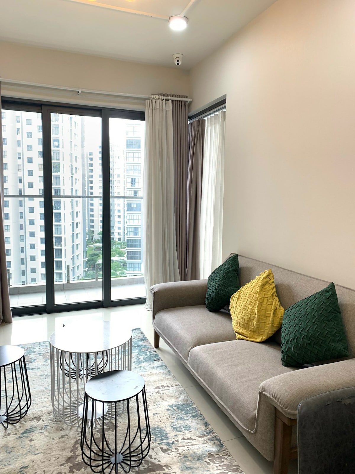 A modern living area features a comfortable sofa adorned with textured pillows, complemented by a light area rug. Three small, round coffee tables sit in front of the sofa, with large windows allowing natural light and offering views of the surrounding buildings.