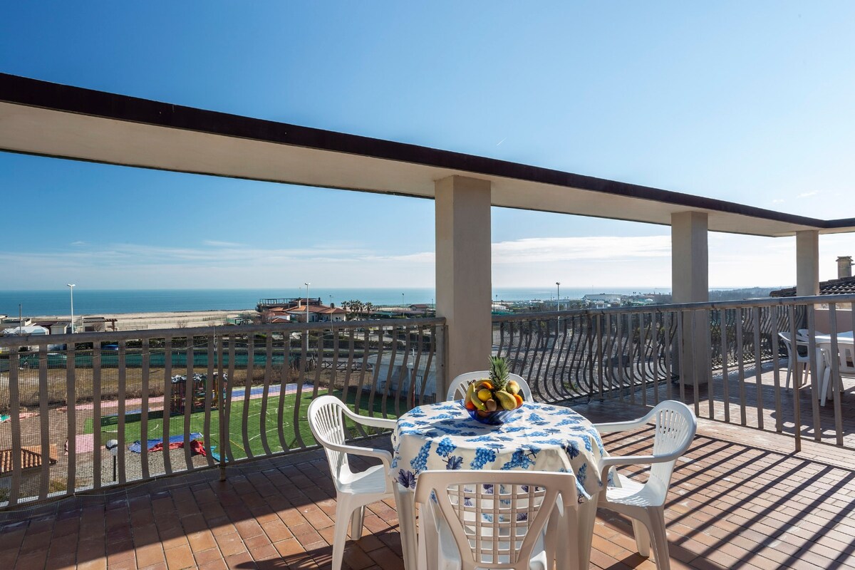 An expansive terrace offers a view of the beach and seafront, featuring a round table adorned with a blue floral tablecloth. Four white plastic chairs surround the table, while the surrounding railing allows for an unobstructed view of the coastline and clear sky.