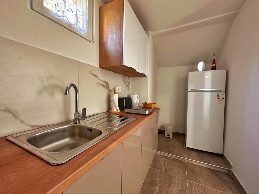 A modern kitchen features sleek cabinetry and a tidy countertop. An undermount sink is positioned next to a stovetop, with a refrigerator visible in the background. Natural light enters through a small window, illuminating the space.