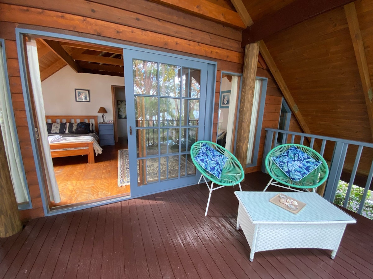 An outdoor seating area features two teal chairs with floral patterns and a small white table. Glass doors open to a cozy bedroom, highlighting wooden beams and warm tones. Natural light fills the space, creating an inviting atmosphere.