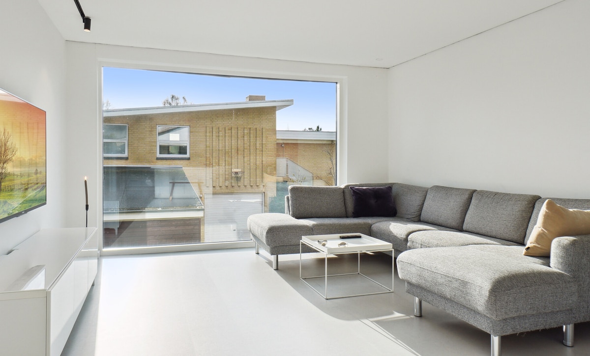 A modern living area is depicted with a spacious gray sectional sofa and a glass coffee table positioned in front of a large window. Natural light fills the room, showcasing a view of the exterior urban landscape. A sleek entertainment unit is situated beneath a wall-mounted television.