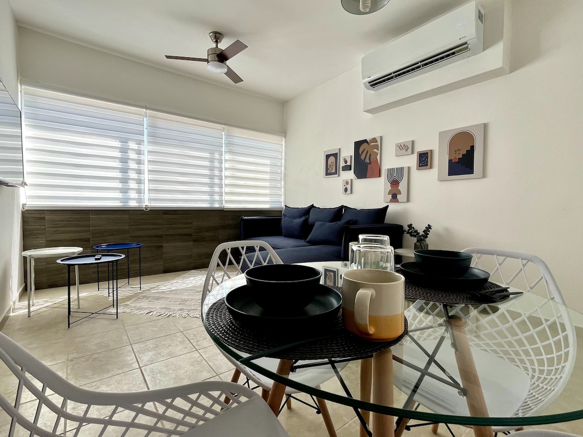 A bright living area features large windows with shades filtering natural light. A dark sofa complements the light walls, while two small tables with black tabletops are positioned nearby. A dining table with white chairs displays black dishware and a neutral-colored cup, creating a casual dining setup.