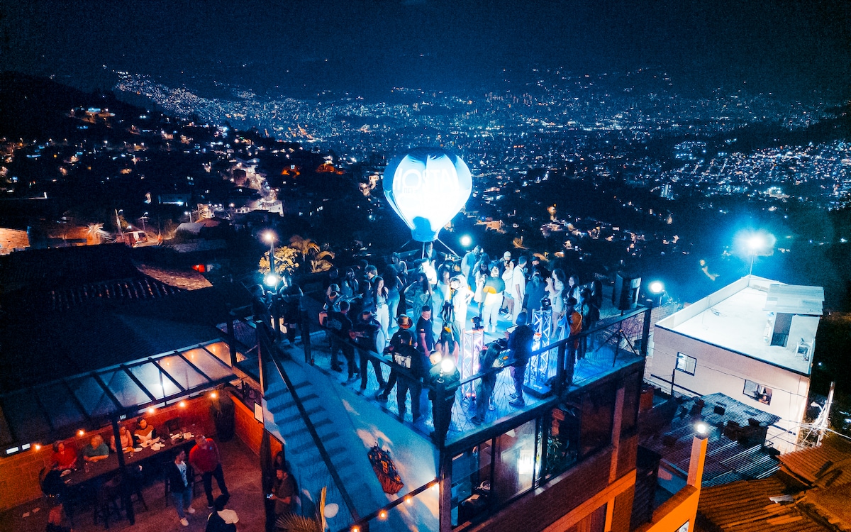 A lively rooftop scene captures a group of people enjoying the evening with the lights of Medellin twinkling in the background. Unique architectural features define the rooftop, with bar areas visible and ample seating spaces. A balloon adds a whimsical touch to the vibrant atmosphere.