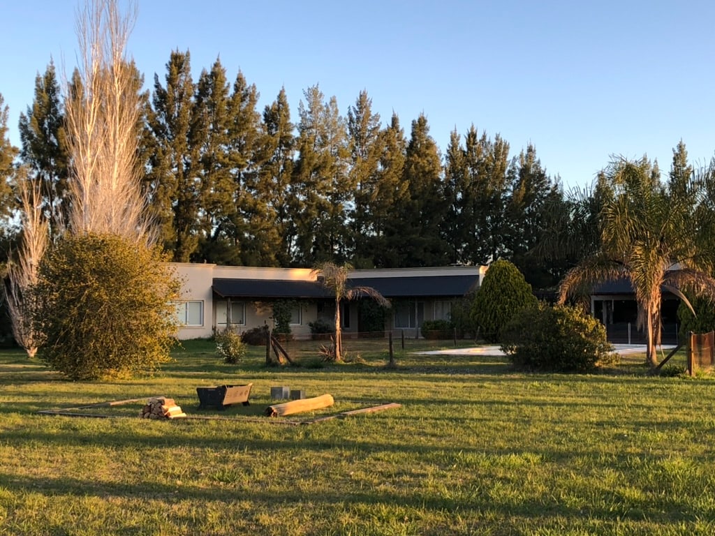 A spacious outdoor setting showcases a residence with a simple façade, accented by palm trees and evergreen foliage. An open area features a fire pit surrounded by logs, set against a backdrop of tall trees under a clear sky.