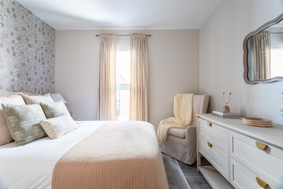 A serene bedroom is furnished with a spacious bed adorned with decorative pillows. Soft curtains frame the windows, allowing natural light to filter in. A cozy armchair is positioned next to a light-colored dresser, enhancing the room's inviting ambiance.