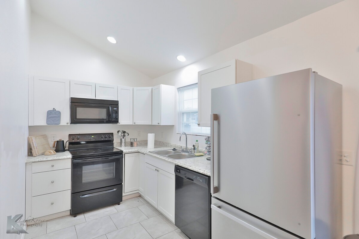 A modern kitchen is shown, featuring white cabinetry, a stainless steel refrigerator, and a sleek black stove. Countertops made of light-colored stone provide ample workspace. Natural light fills the room through a nearby window above the sink, enhancing the bright, airy atmosphere.