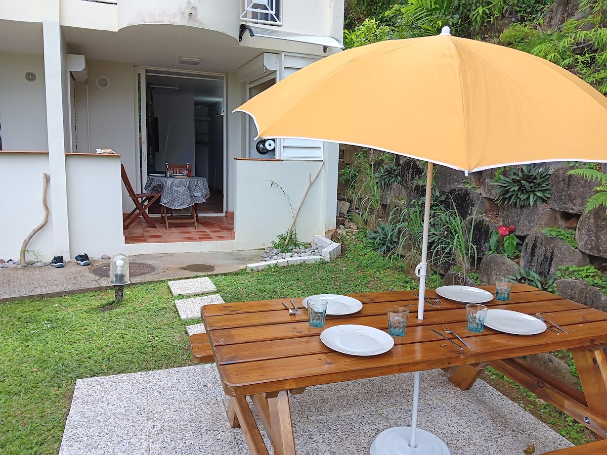 An outdoor dining area is presented with a wooden table set for four, accompanied by matching chairs and a large yellow umbrella. A well-maintained garden surrounds the space, and a path leads to an entrance featuring a partially visible interior area.