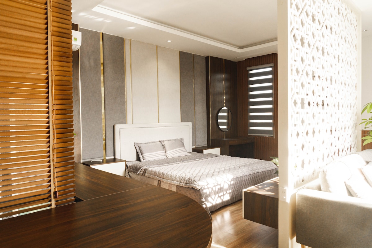 A spacious bedroom features a large bed with a quilted cover. Natural light fills the room through window blinds, highlighting the textured walls and wooden accents. A circular mirror is positioned on the wall, while a comfortable seating area is present in the corner.