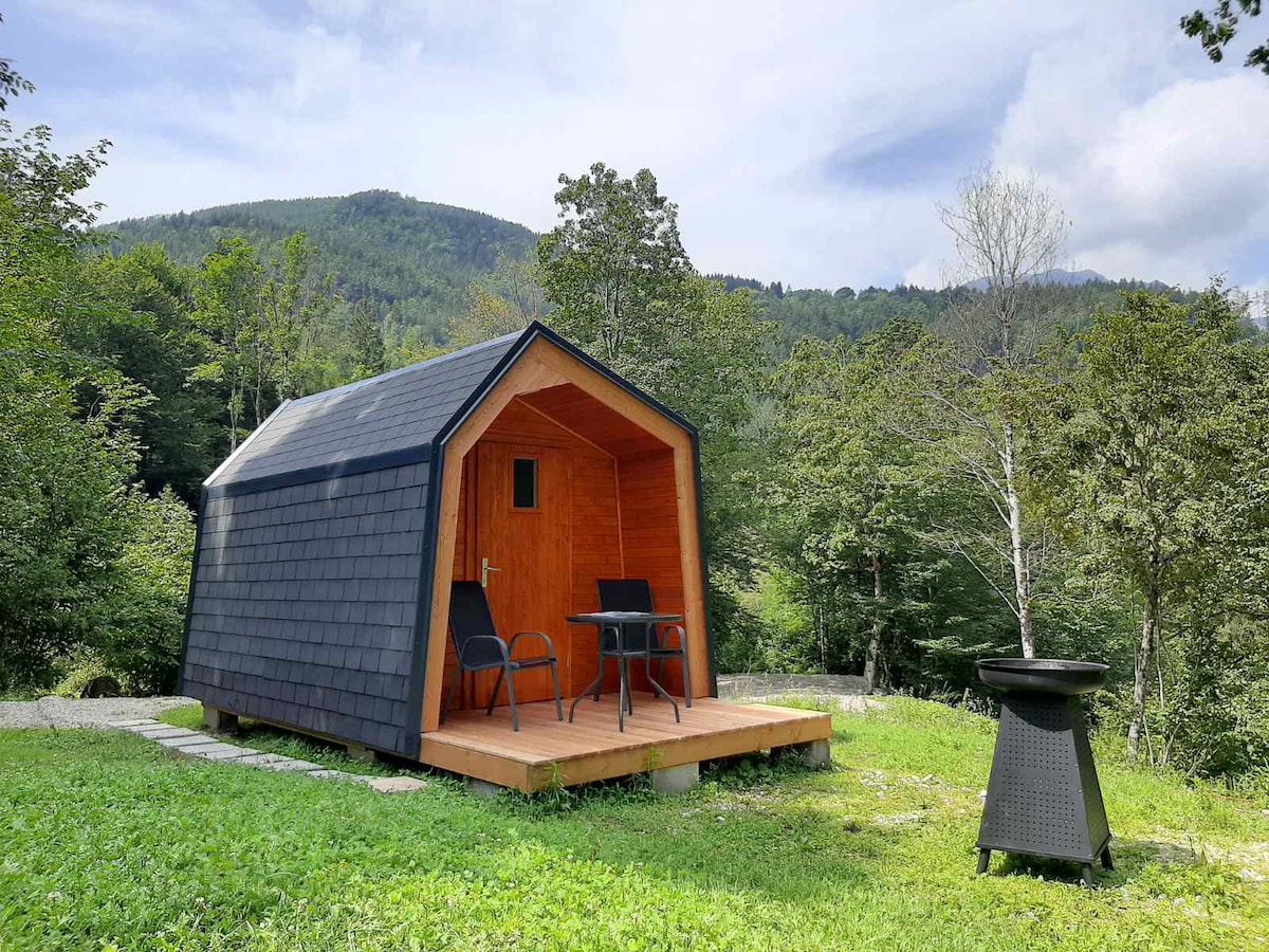 A cozy glamping cabin is featured, set among lush greenery. The cabin has a distinctive a-frame design with wooden accents and a covered porch. A small outdoor seating area with a table and two chairs is visible, along with a fire pit placed nearby.