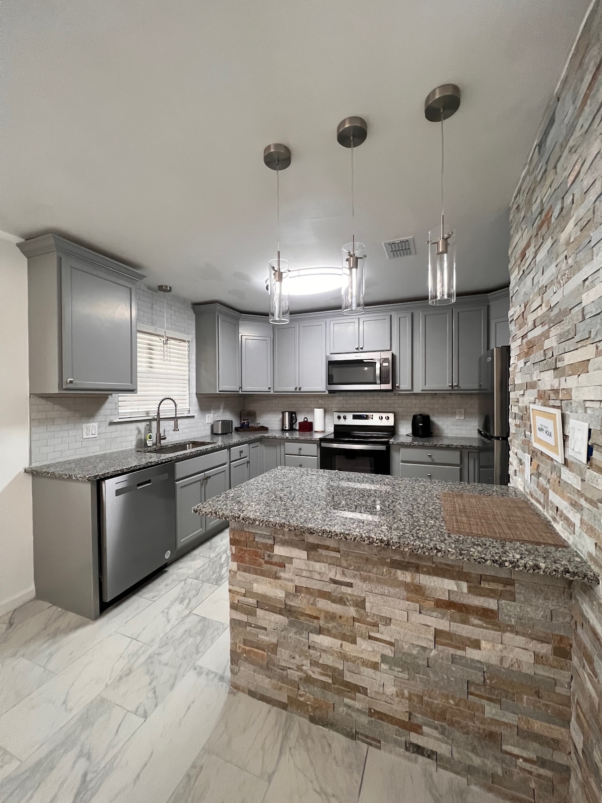A modern kitchen is highlighted by gray cabinetry and a granite countertop. Stainless steel appliances, including a microwave and refrigerator, are neatly arranged. Natural light filters in through a window, and elegant pendant lighting is suspended above the kitchen island crafted from textured stone.