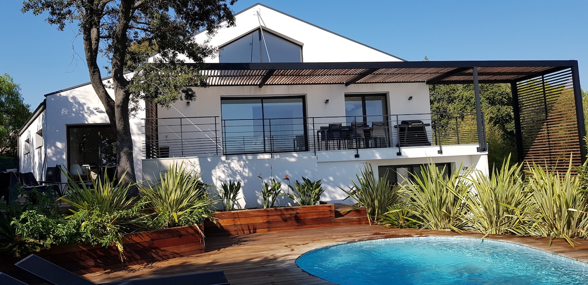 The exterior of the villa is highlighted, featuring a modern design with large windows and a spacious terrace. A wooden deck surrounds a circular pool, complemented by tropical plants and outdoor furniture, offering a serene atmosphere in a sunny setting.