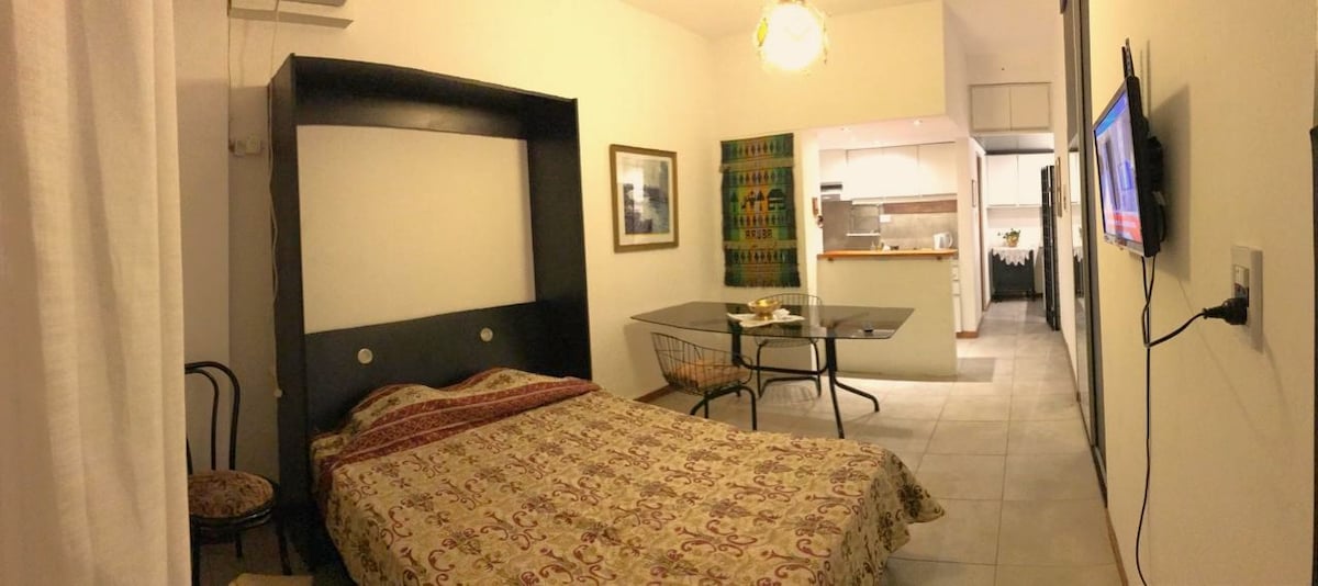 A cozy studio apartment is presented, featuring a spacious bed with a patterned bedspread. A small table with chairs is positioned near the kitchenette, enhancing functional living space. Light walls and simple furnishings contribute to an inviting atmosphere, while a television is mounted on the wall.