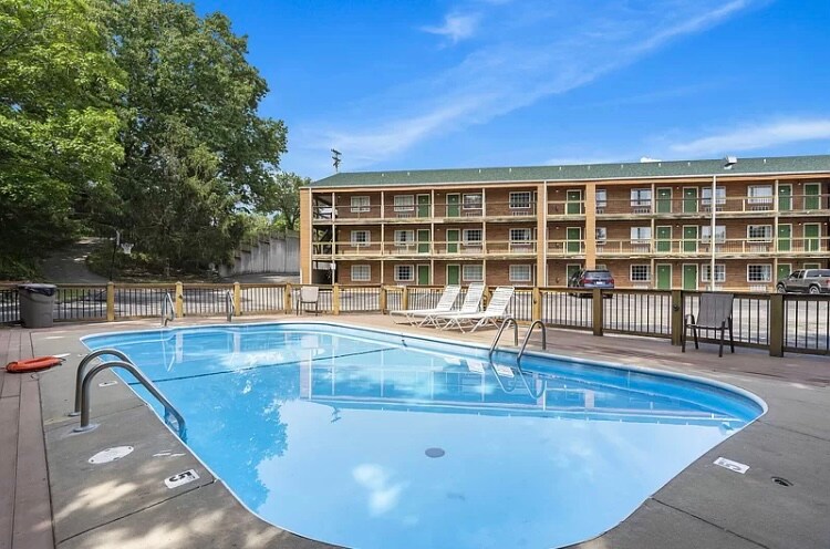 An outdoor swimming pool is visible, featuring a smooth blue surface and steps leading into the water. White lounge chairs are neatly arranged around the pool area. In the background, a three-story building with multiple green doors is present, surrounded by lush greenery.