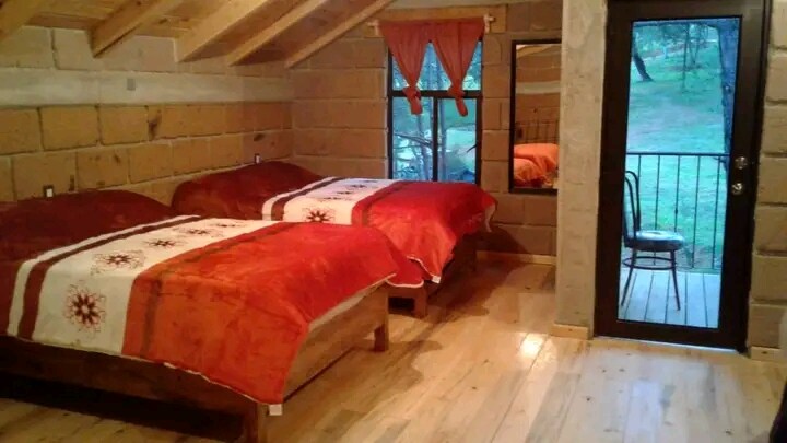 The inviting sleeping area features two spacious beds adorned with vibrant red covers. Natural light filters through a window, illuminating the wood-paneled walls and polished wooden floor. A mirror and a small chair complement the cozy decor, while a door leads to the outside.