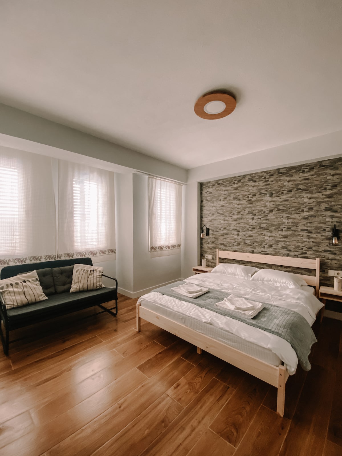 A bright room features a spacious bed with neatly arranged linens, complemented by a cozy sofa. Natural light streams through window coverings, highlighting the textured stone accent wall. Warm wood flooring adds a welcoming touch, creating a calming environment.