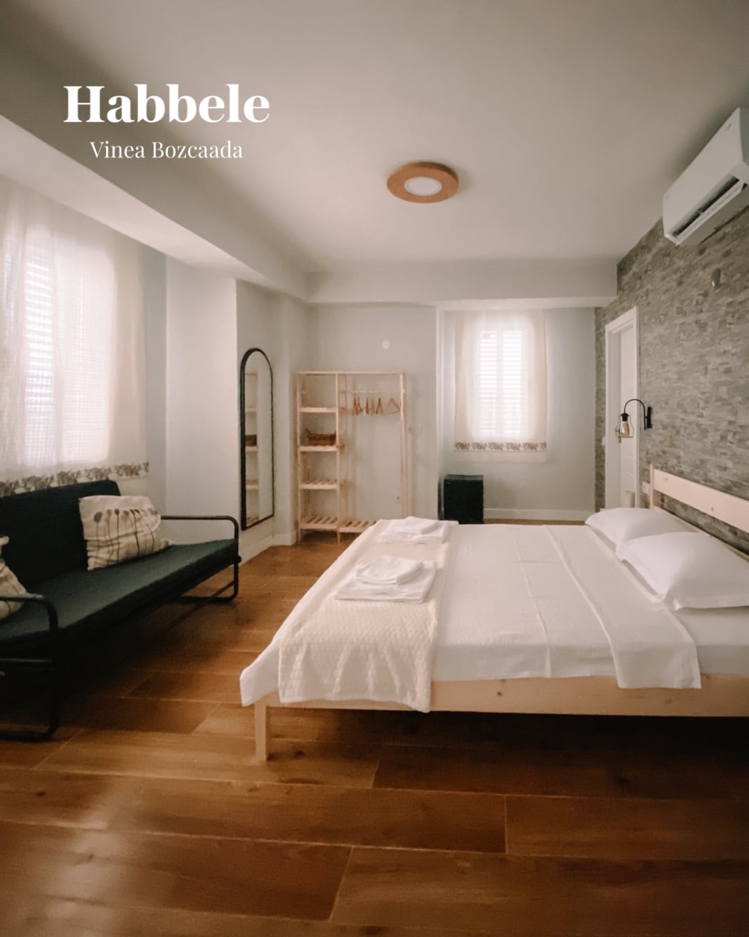 A spacious room features a large double bed and a convertible sofa that accommodates up to four guests. Natural light enters through the window, illuminating the wooden floor. A mirror and clothing rack are positioned against the wall, contributing to the room's practical layout.