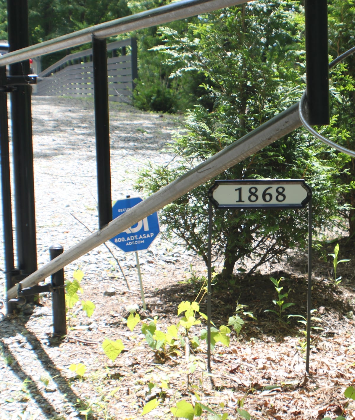 A pathway is visible with a black and white house number sign displaying '1868'. Nearby, a security sign is placed near greenery. The surrounding area features gravel and small plants, suggesting a natural setting.