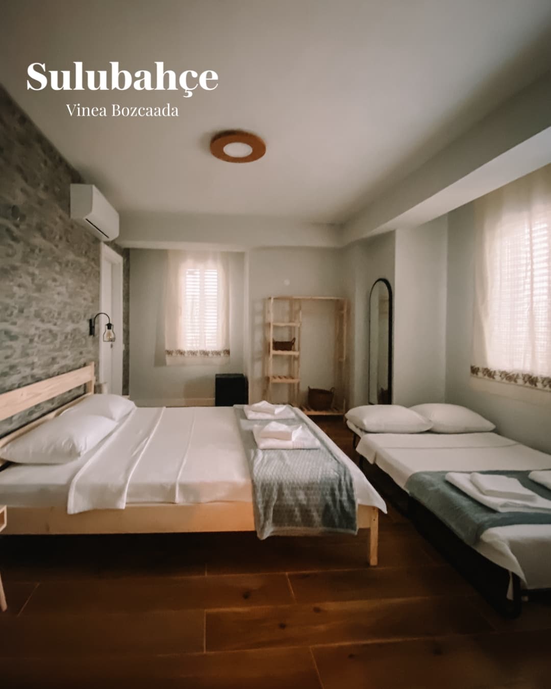 The room features a spacious double bed alongside a convertible sofa bed, accommodating up to four guests. Natural light filters through sheer curtains, enhancing the serene atmosphere. A wardrobe and a mirror are positioned against the wall, contributing to the room's functionality.