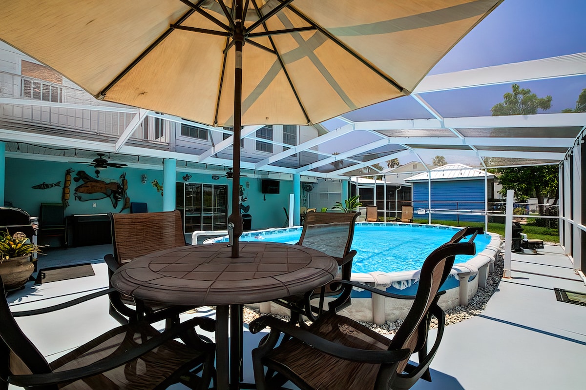 A spacious patio area surrounds the heated pool, featuring a table with an umbrella and several comfortable chairs. The screened enclosure allows for unobstructed views of the clear blue water and is complemented by a grill station nearby.