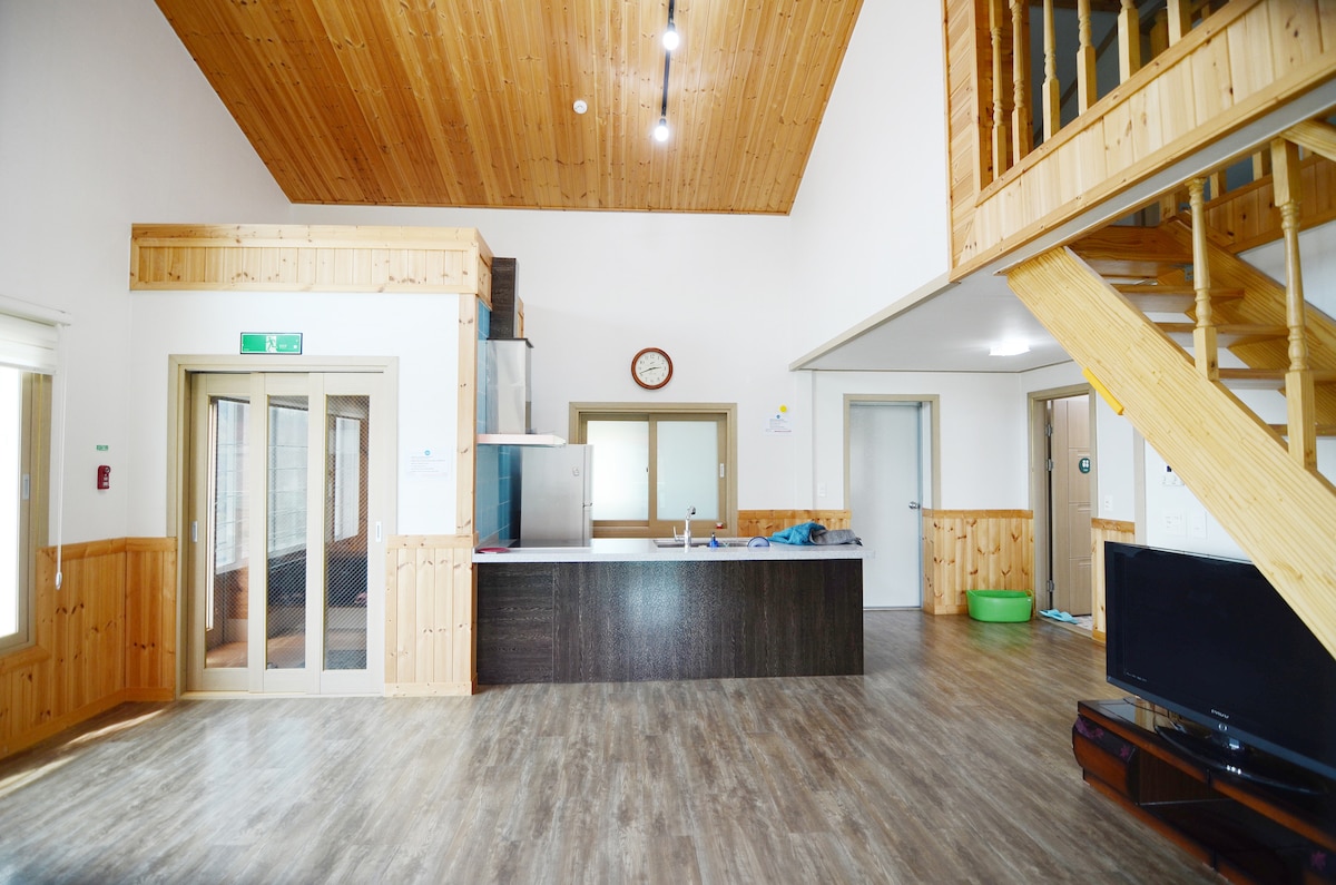 The image displays an open living area featuring high ceilings with wooden beams. A spacious kitchen is visible on one side, equipped with modern appliances. Large windows provide natural light, while a wooden staircase leads to the upper level, enhancing the area's airy feel.