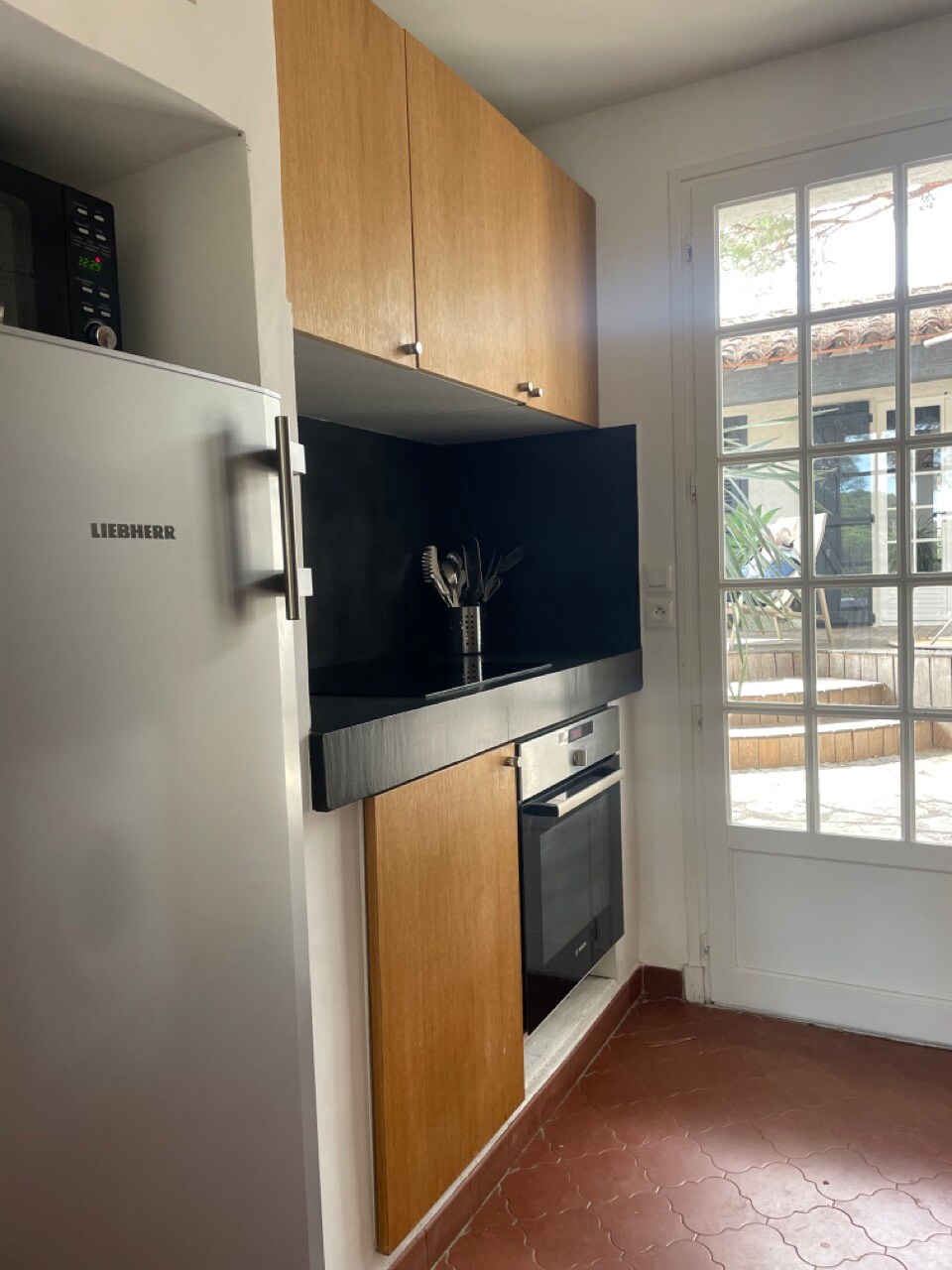 A compact kitchen is shown, featuring wooden cabinets and black countertops. A refrigerator and an oven are visible, along with kitchen utensils arranged on the counter. Natural light enters through a glass door, which opens to the patio, enhancing the bright and functional space.