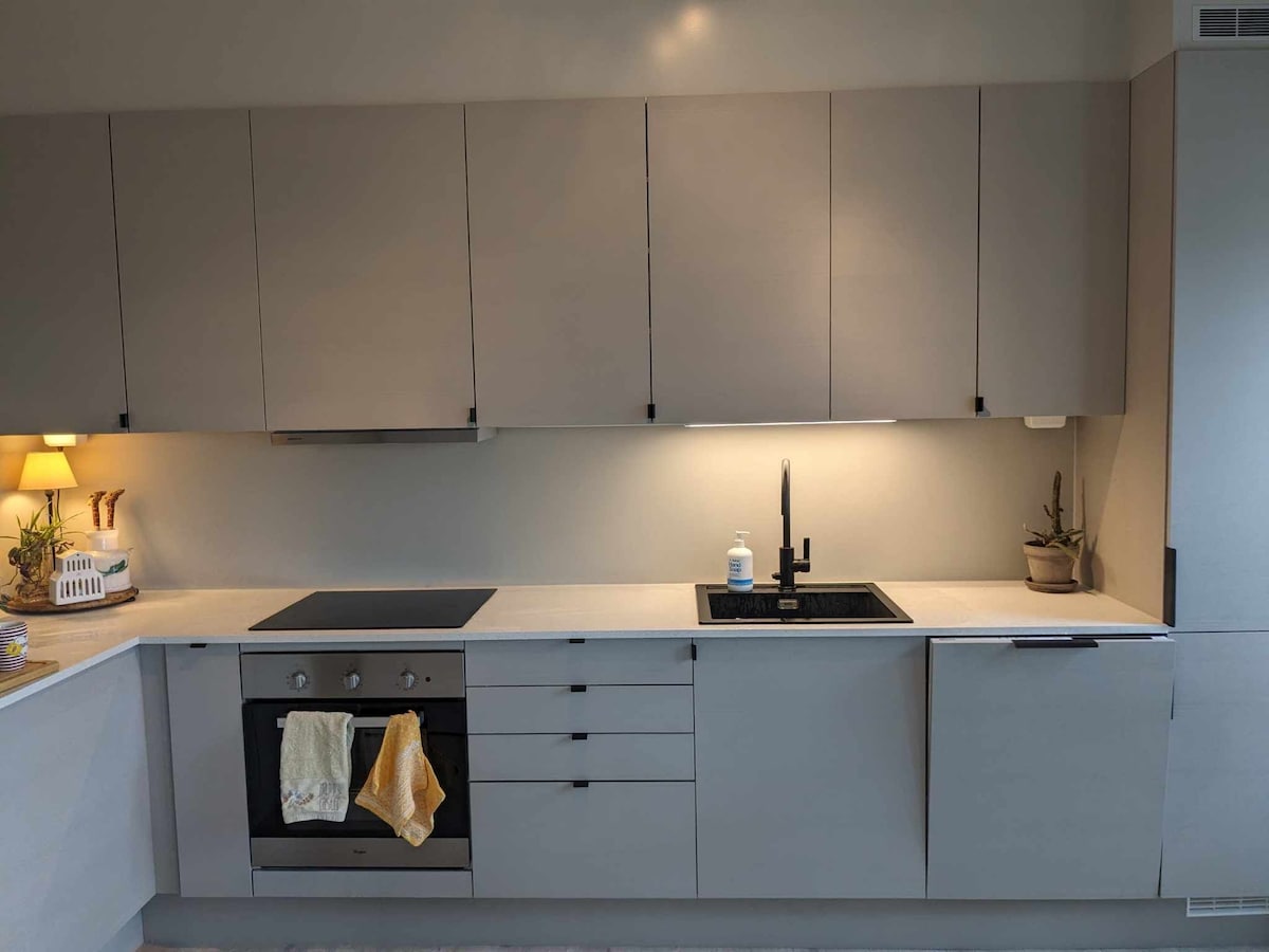 A modern kitchen is depicted with sleek, light grey cabinetry and a smooth countertop. Essential appliances are integrated, including an oven and a black sink. Soft lighting enhances the workspace, complemented by potted plants and a lamp providing a gentle glow.