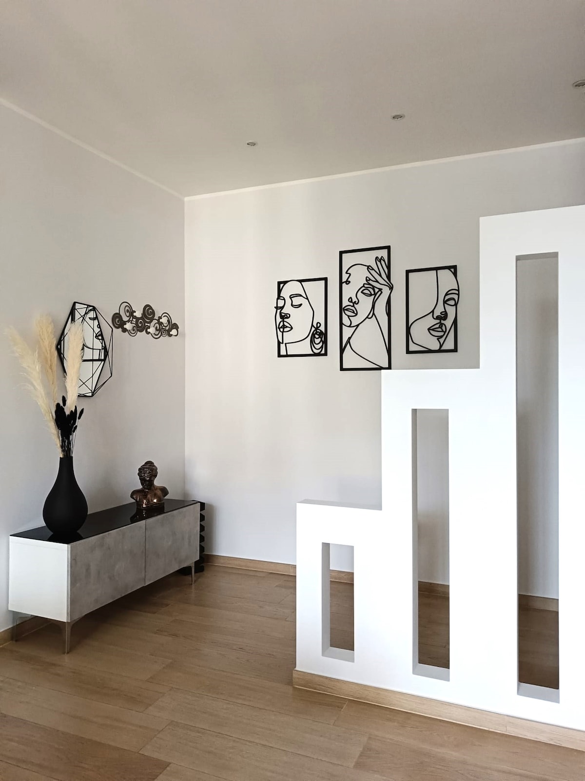 A stylish entryway features minimalist wall art depicting abstract faces. A modern console table with a glossy finish and a decorative vase adds sophistication. Natural wood flooring complements the clean lines of the architectural design, creating a warm ambiance.