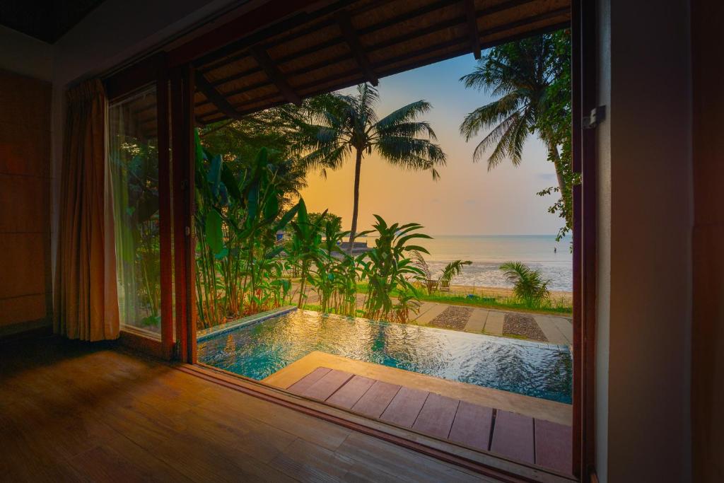 A serene view of a private pool is framed by large glass doors, open to lush greenery and the ocean beyond. The horizon is softly illuminated by sunset hues, with palm trees swaying gently in the breeze.