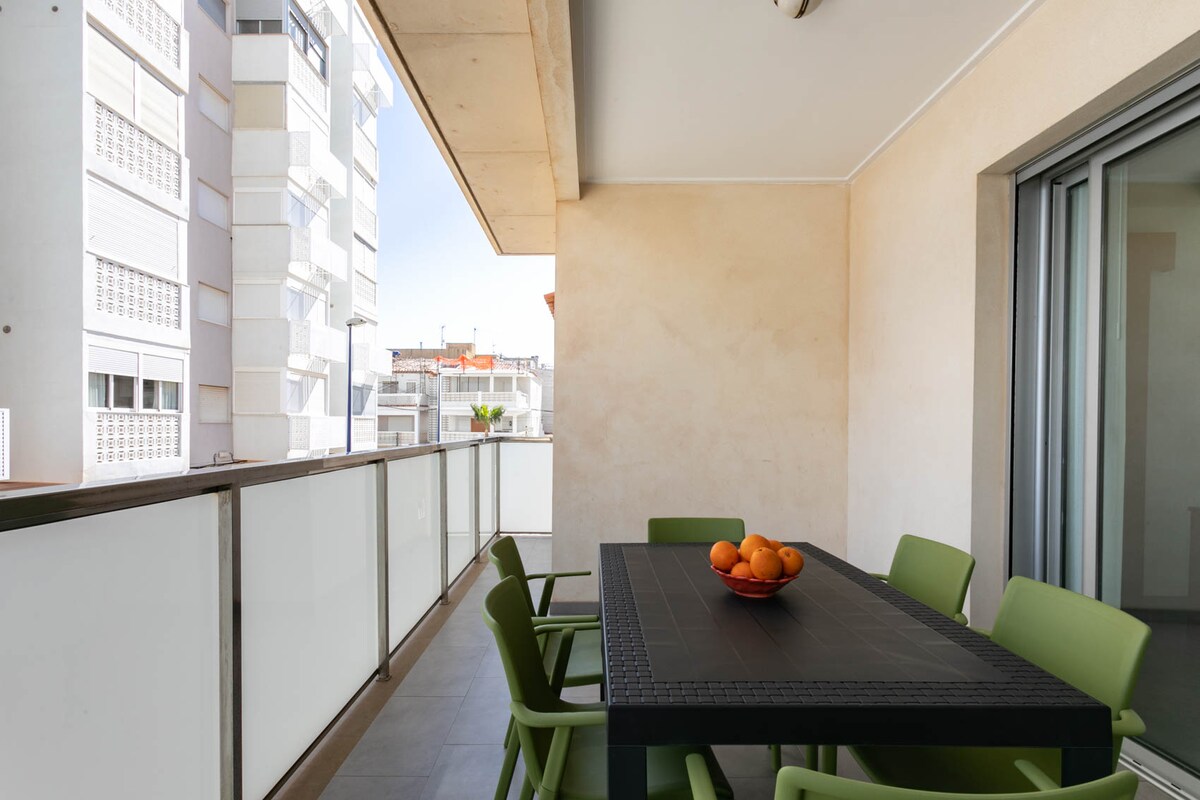 A balcony is presented with a modern table surrounded by green chairs. A bowl of oranges sits at the center of the table. The balcony is enclosed by clear glass railings, providing a view of surrounding buildings and allowing natural light to enter the space.