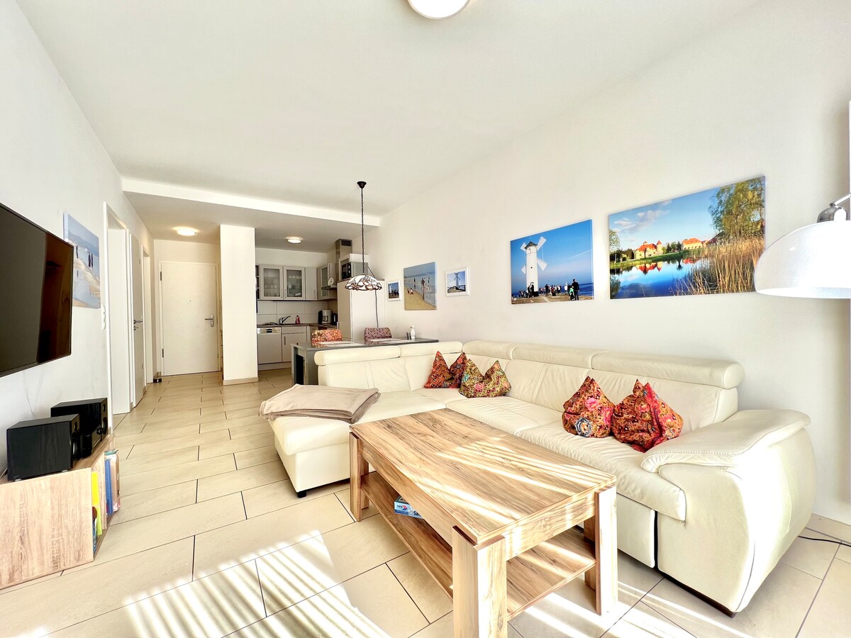 A spacious living area features a large white sofa adorned with colorful cushions. A wooden coffee table sits in the center, complemented by a mounted flat-screen TV. Bright artwork decorates the walls, and natural light fills the room through a nearby window.