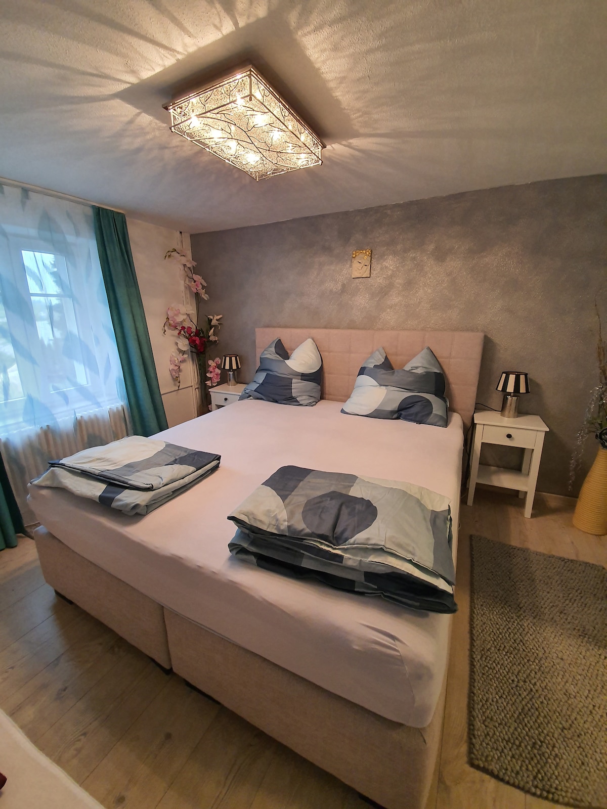 A private bedroom features a spacious bed adorned with patterned bedding and decorative pillows. Natural light filters through sheer curtains, illuminating the room's soft tones. A sleek nightstand and a charming light fixture enhance the overall design.