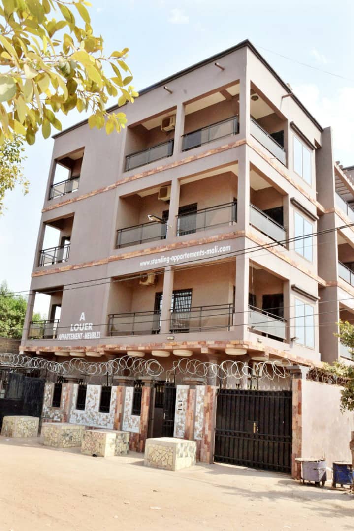 A modern three-story building is depicted, featuring multiple balconies with black railings. The exterior is painted in a neutral color, framed by greenery. A secure entrance is visible at the bottom, showcasing a decorative wall and barbed wire above the gate.