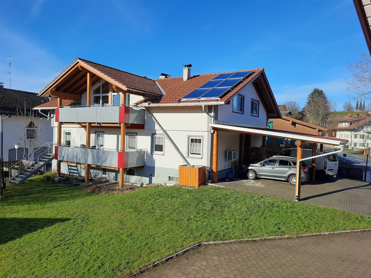 The exterior of a two-story building is shown, featuring a terracotta roof and solar panels. The structure is surrounded by a grassy area and includes an adjacent driveway with parked cars. A porch area with wooden beams is visible, enhancing the overall inviting appearance.