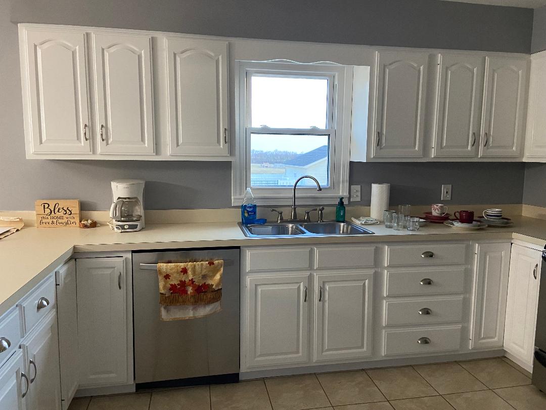 A fully equipped kitchen is shown featuring white cabinetry and a neutral countertop. Stainless steel appliances are visible, including a refrigerator and dishwasher. A window above the sink offers views of the exterior. Light-colored tiles cover the floor, creating a clean and functional space.