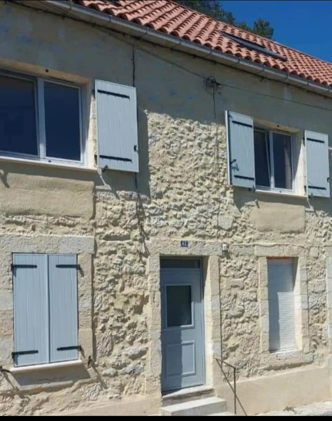 The exterior of the town house features traditional stone walls with a light beige finish. Windows are adorned with blue shutters, some partially closed. The entrance is marked by a blue door, leading to the cozy interior. A simple staircase provides access to the front door.