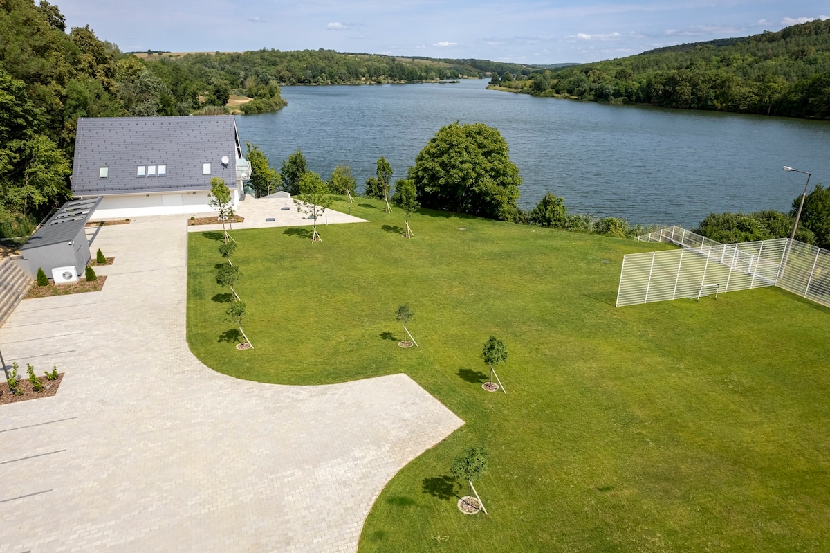 An expansive lawn extends next to a serene water body, bordered by trees. The modern house features a dark roof and a spacious driveway made of stone. The surroundings are peaceful, with no visible neighboring structures, promoting a sense of privacy and connection with nature.