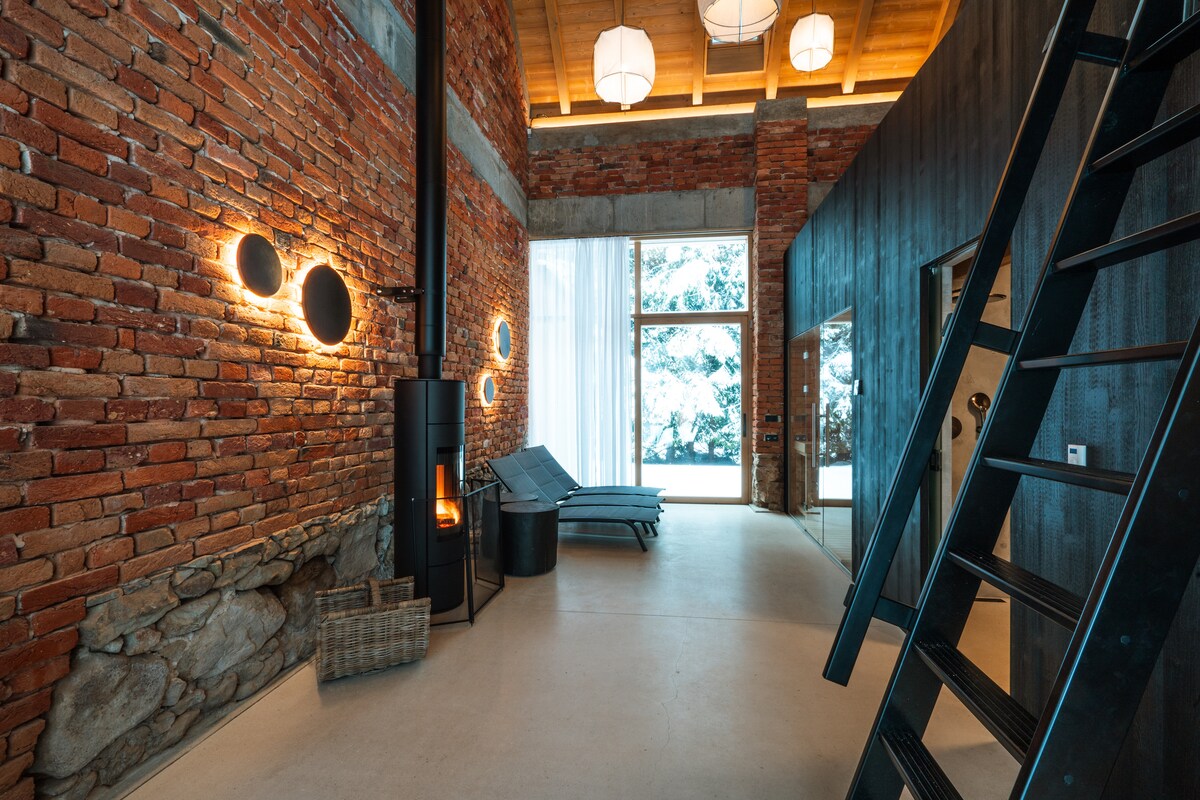 An industrial loft space features exposed brick walls and modern light fixtures. A sleek wood-burning stove stands near a stone wall, while two lounge chairs are positioned nearby. Large glass doors reveal a snow-covered outdoor view, enhancing the serene atmosphere.