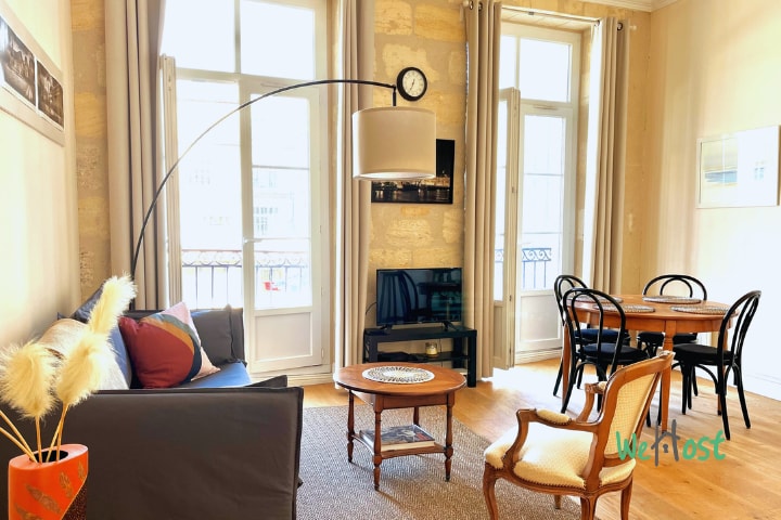 A spacious living area is featured, showcasing tall windows that invite natural light. The room comprises a comfortable sofa, a decorative armchair, and a wooden dining table set for four. A floor lamp provides additional illumination, enhancing the inviting atmosphere.