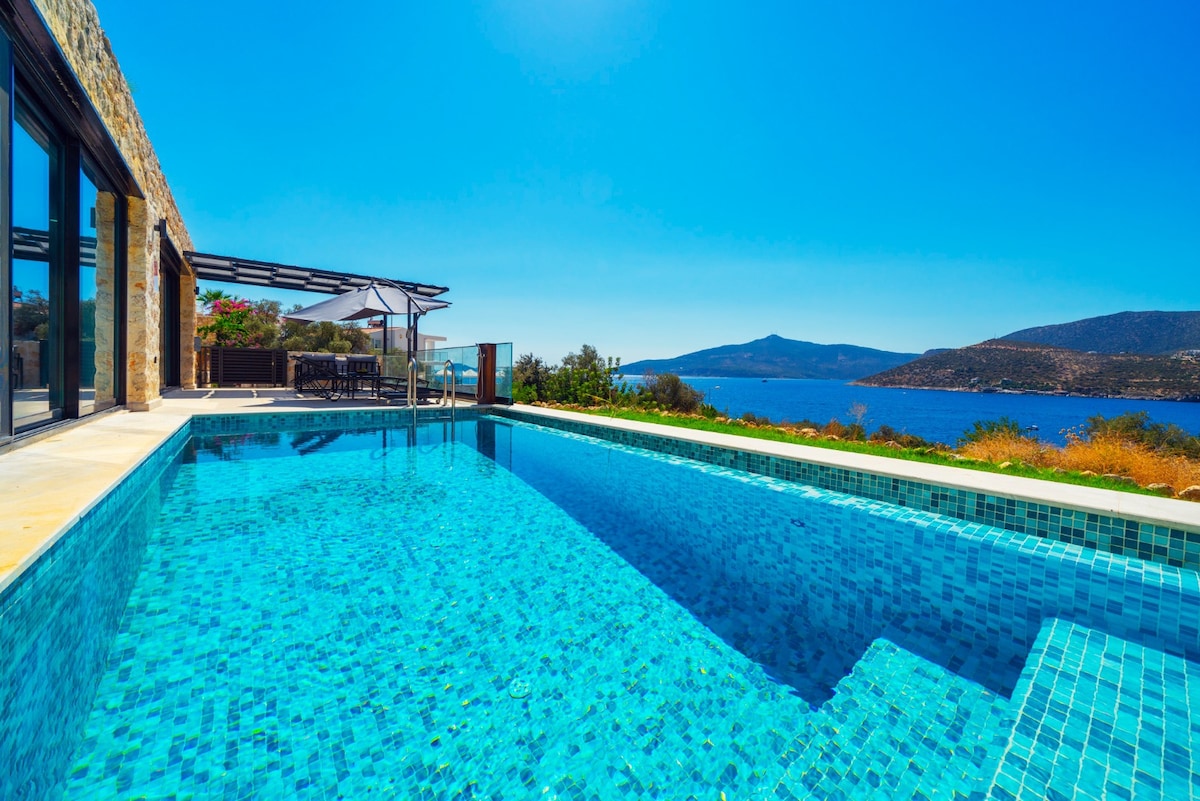 A crystal-clear infinity pool extends towards a panoramic view of the sea, with sunlight reflecting off the water's surface. Stone walls complement the surrounding landscape, while outdoor seating is provided under a shaded pergola, creating a perfect spot for relaxation.