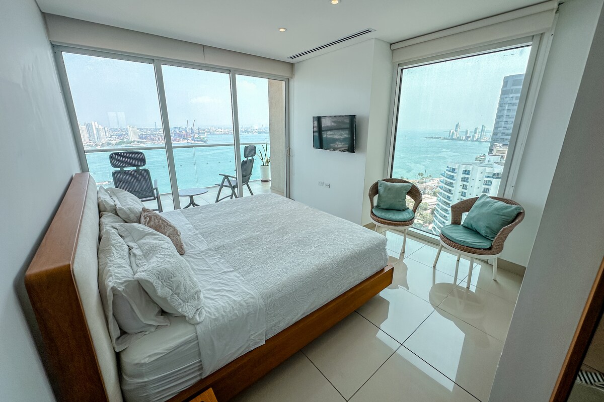 A spacious bedroom features a large bed dressed in white linens. Floor-to-ceiling windows offer views of the ocean and cityscape. Two stylish chairs are positioned beside the bed, with a flat-screen TV mounted on the wall opposite. Natural light fills the room.