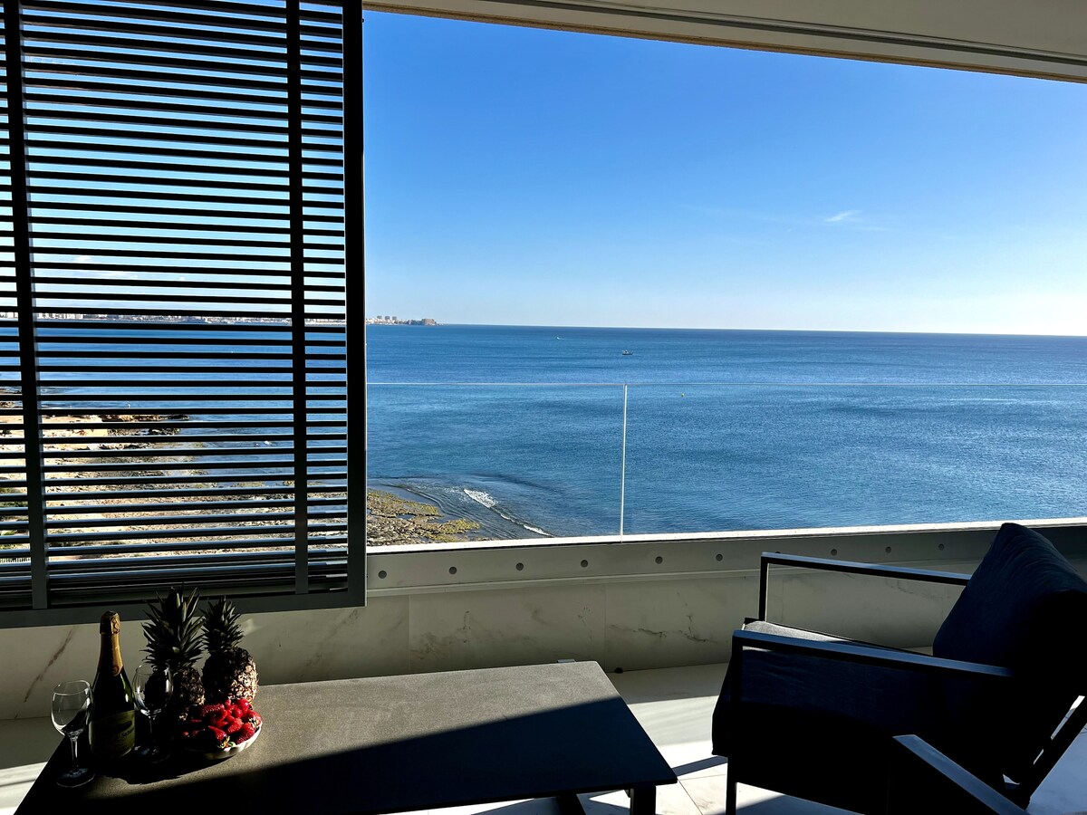 A spacious terrace reveals expansive sea views, framed by large sliding doors. A sleek coffee table displays fruits and drinks, while a comfortable chair invites relaxation. The serene blue waters and clear sky enhance the coastal ambiance.